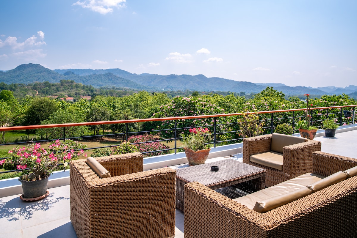 A spacious outdoor terrace is furnished with comfortable wicker seating and a low table, offering views of lush greenery and rolling hills in the distance. Potted plants enhance the natural setting, while the bright blue sky completes the inviting atmosphere.