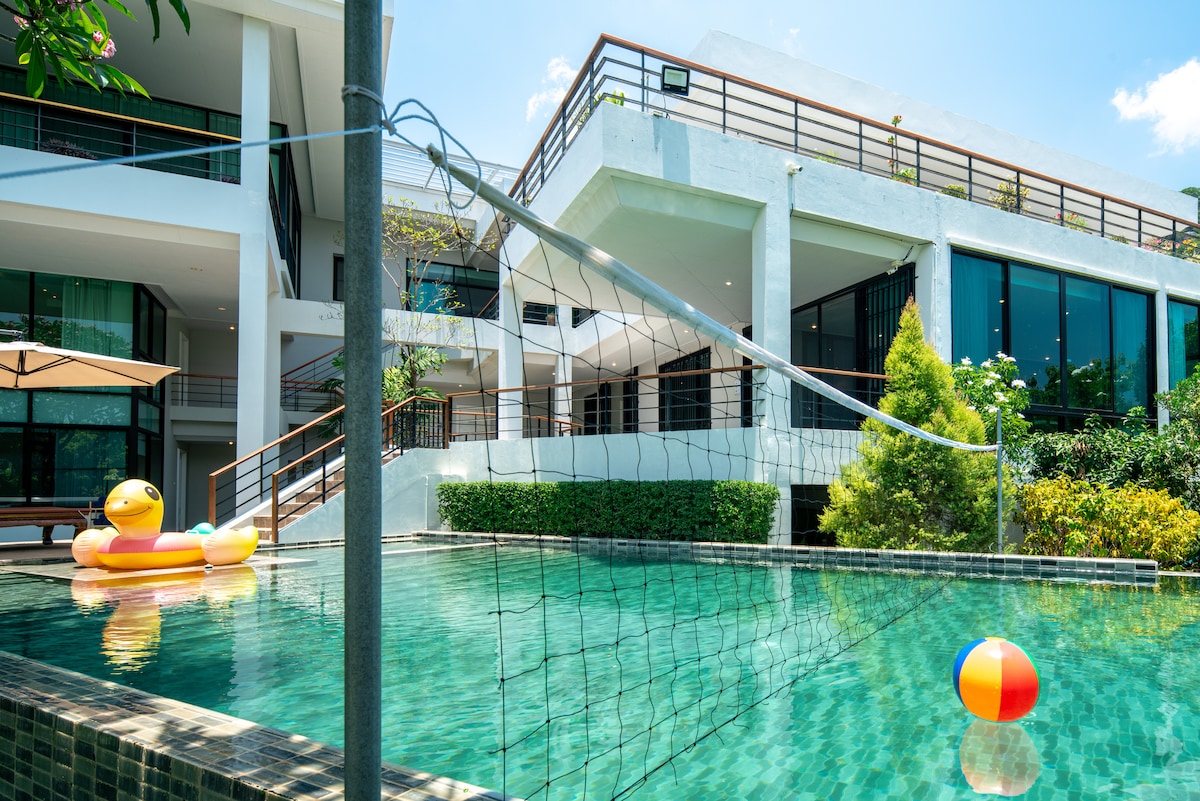 A serene swimming pool is framed by modern architecture, featuring a volleyball net and colorful inflatable toys floating on the water. Lush greenery surrounds the pool, enhancing the tranquil ambiance of the outdoor space.