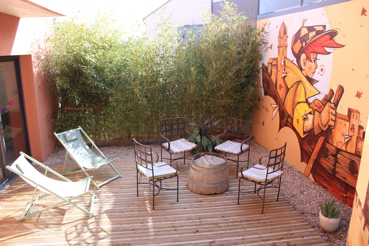An outdoor terrace features a wooden deck surrounded by bamboo for privacy. Two lounge chairs and four metal chairs with cushions are arranged around a barrel table. A colorful mural adds a vibrant touch to the space, enhancing the relaxed atmosphere.
