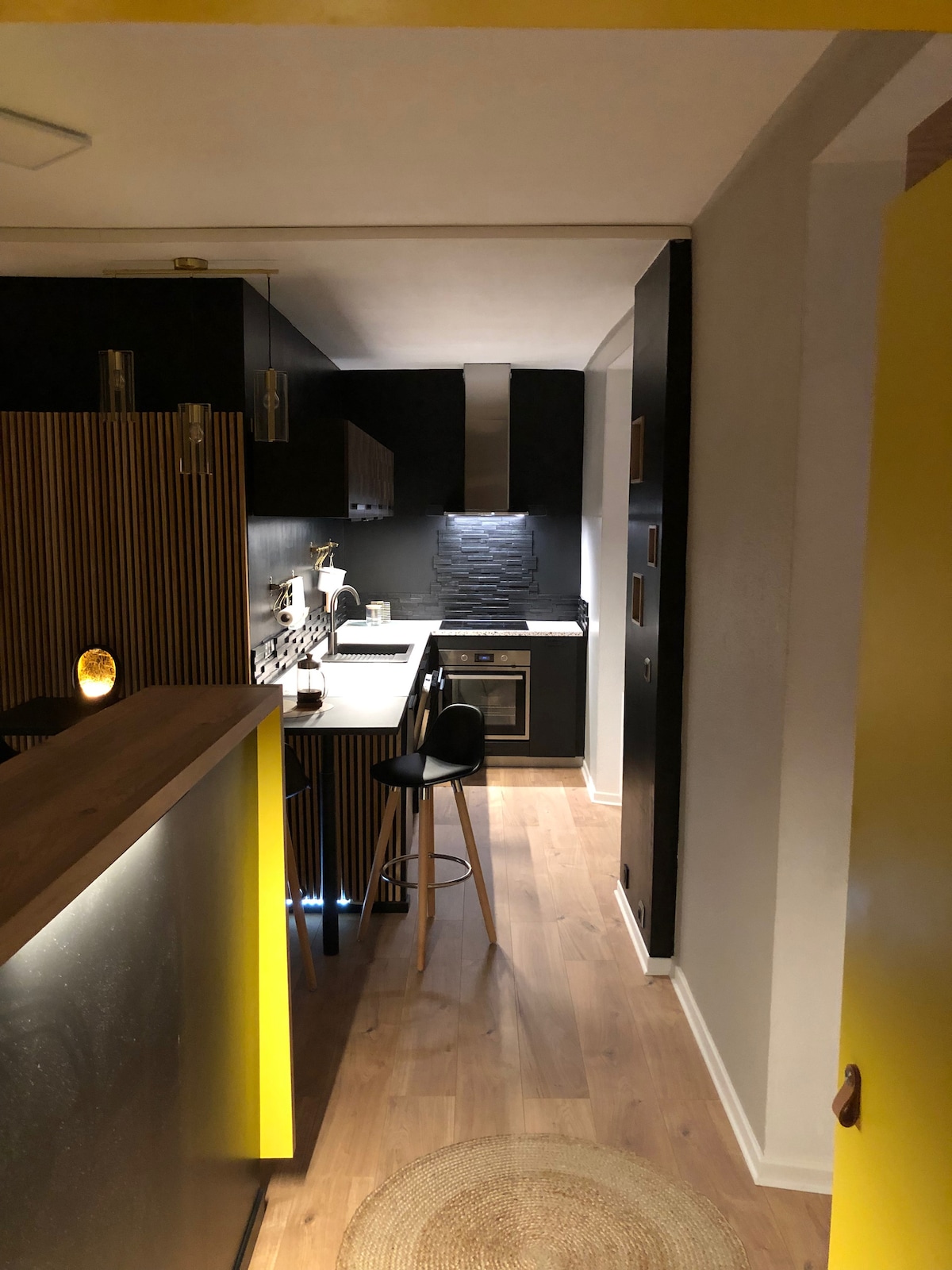 The kitchen area is designed with modern cabinetry, featuring a combination of dark and light tones. A small bar with stools is situated near the entrance, while a sleek stove and sink are visible in the background, illuminated by warm lighting.