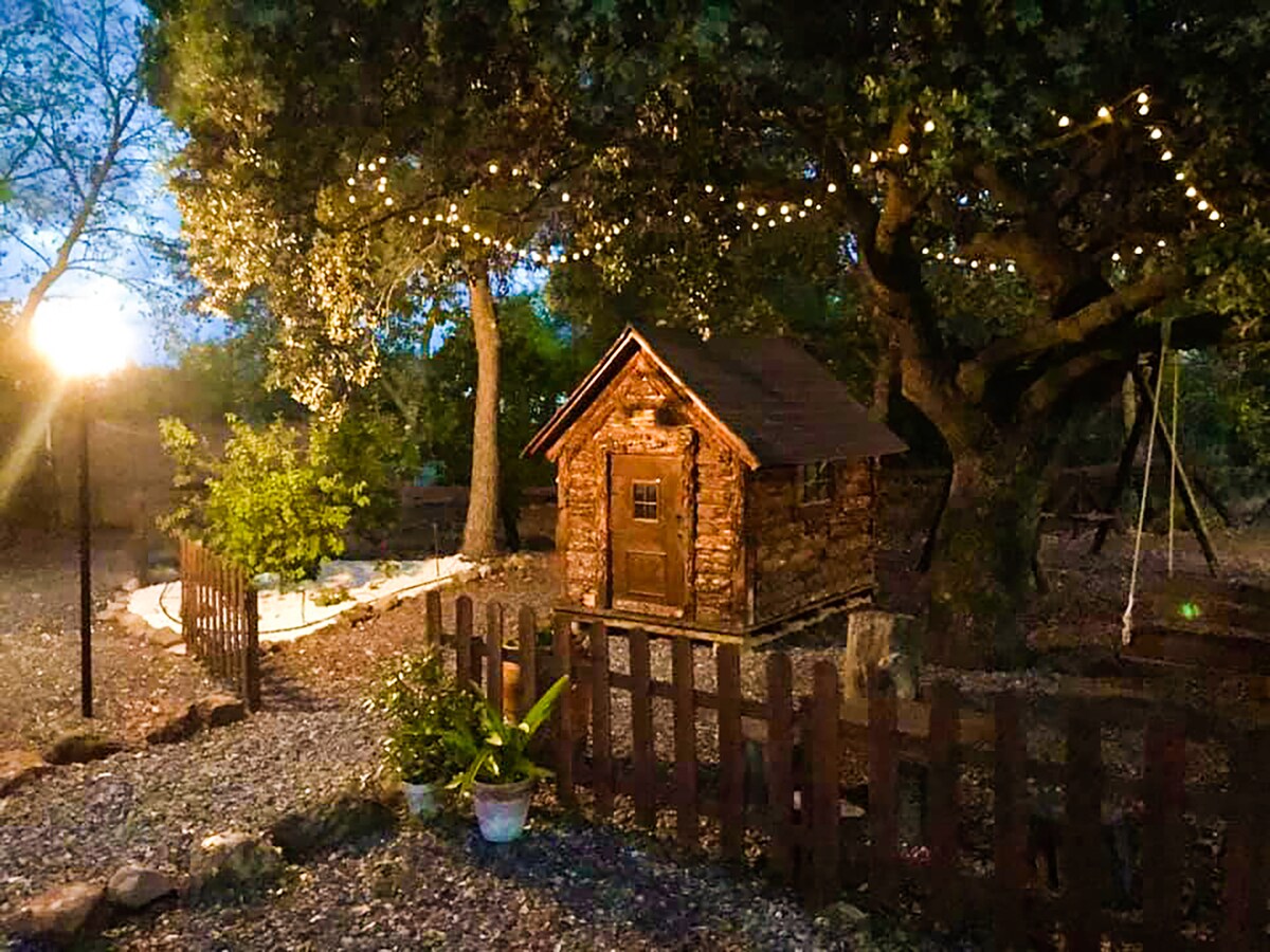 A quaint wooden cabin is nestled among trees, illuminated by soft string lights. The surrounding area features a gravel pathway and a wooden fence. A small garden with potted plants adds greenery, while a swing hangs from a sturdy branch nearby.