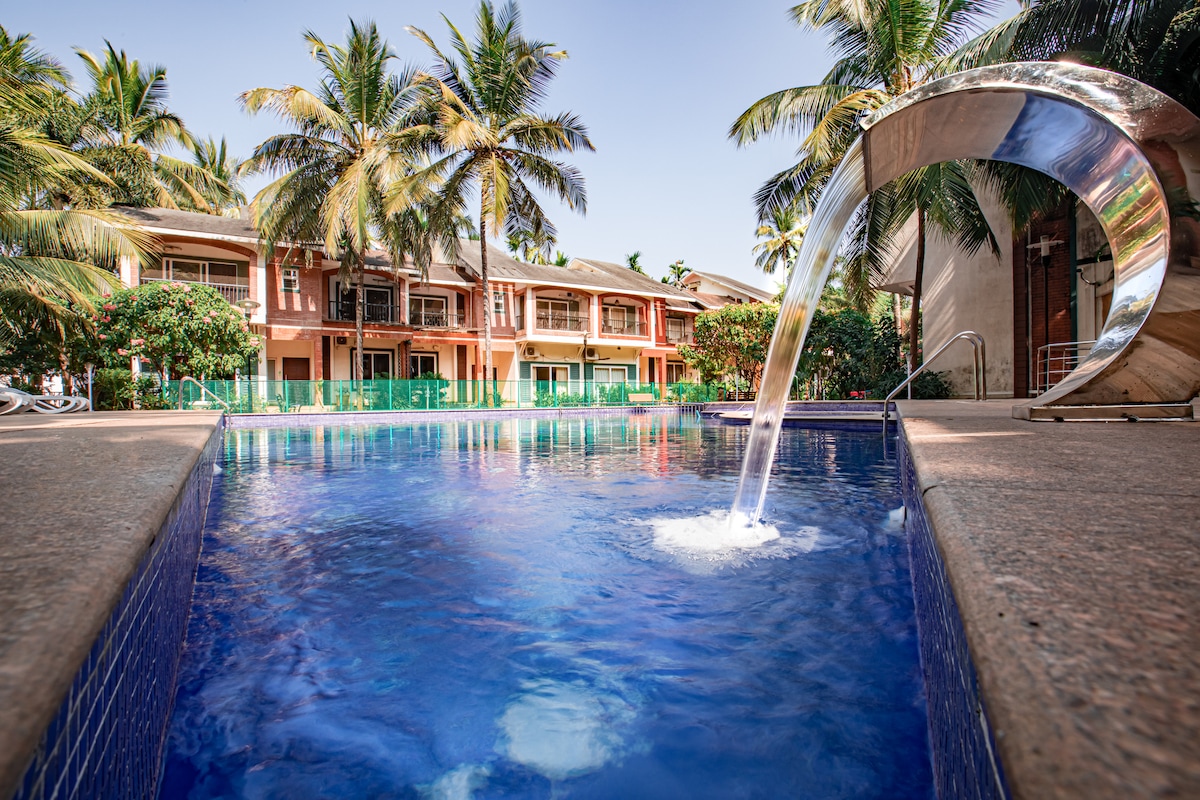 A serene pool is featured, with clear blue water reflecting the sky. The pool's edge is surrounded by lush palm trees and well-maintained greenery. In the background, a two-story villa with balconies is visible, offering a comfortable retreat.