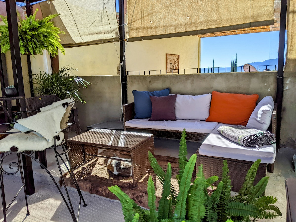 A comfortable rooftop patio features a shaded seating area with a cushioned couch adorned with colorful pillows. A coffee table rests in front, complemented by potted ferns and a decorative piece on the wall. Panoramic views of the nearby mountains are visible in the background.