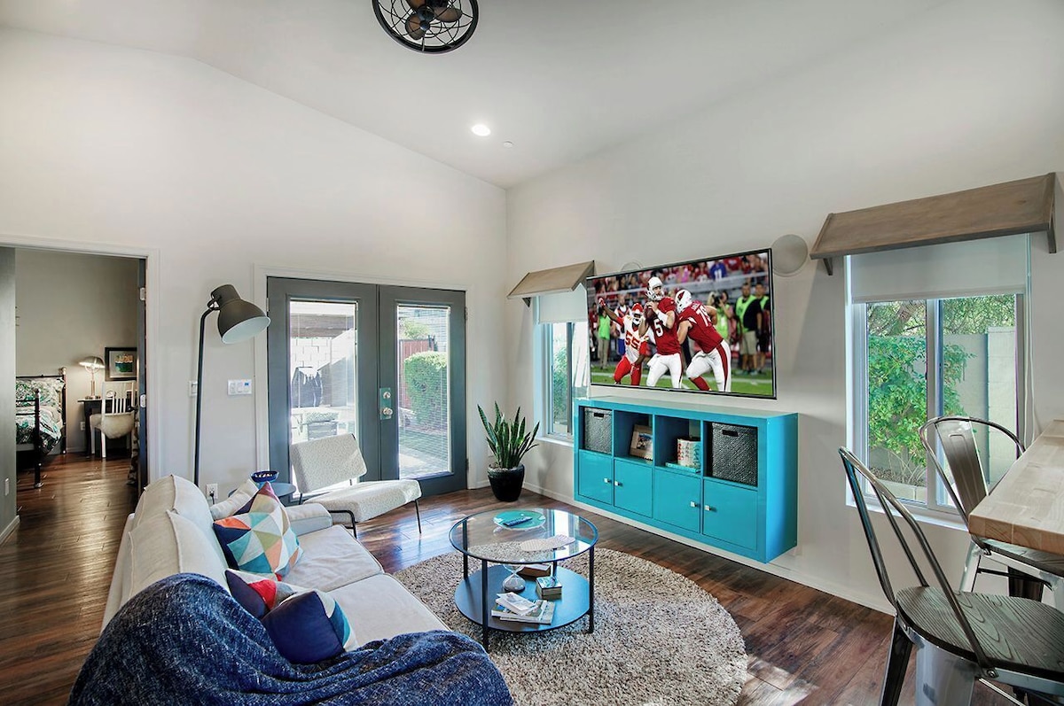 A spacious living area features a large television mounted on a light-colored wall, displaying an athletic event. A circular glass coffee table is centered on a textured rug, surrounded by a comfortable sectional sofa with decorative pillows. Natural light enters through multiple windows with minimalist treatments.