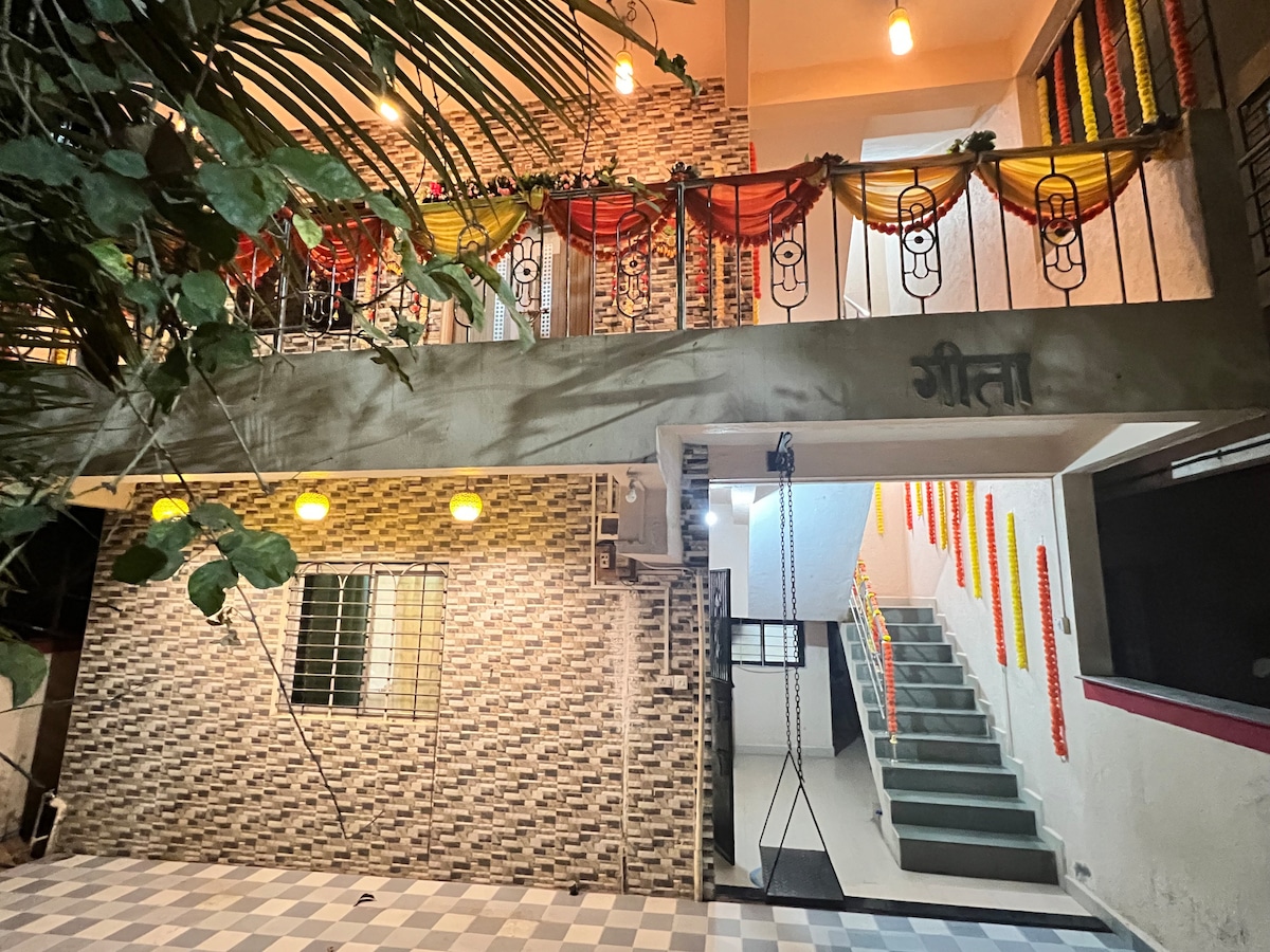 The entrance to the bungalow features a welcoming façade adorned with colorful decorations. The stairway, flanked by a railing, leads to the upper level. Soft lighting enhances the inviting atmosphere, with greenery partially visible, providing a warm and cozy entry point.