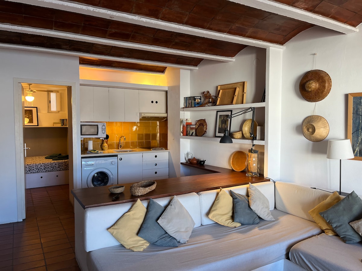 An open concept living space features a comfortable seating area adorned with cushions in various colors. A kitchen with modern appliances, including a washing machine, is visible in the background. Decorative items and artwork are arranged on shelves, contributing to the room's character.