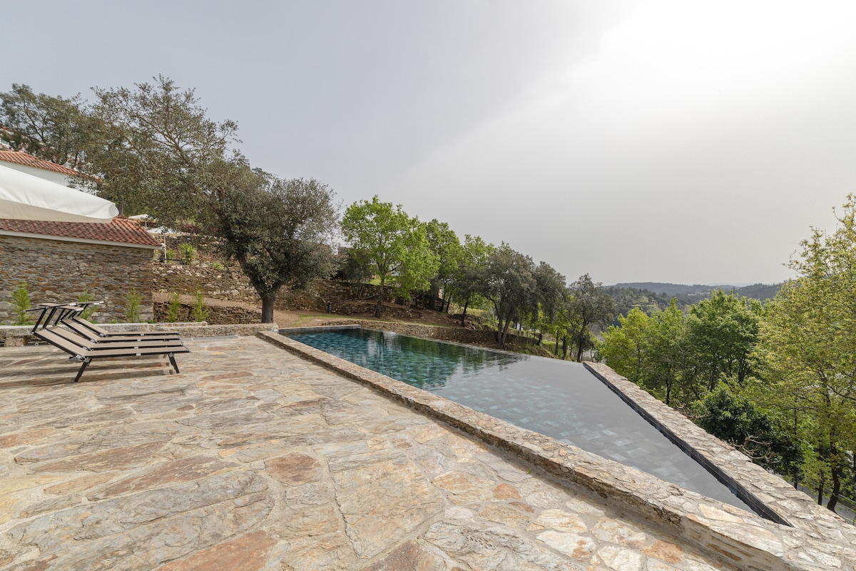 An outdoor swimming pool with a natural stone surround reflects the lush greenery of the surrounding landscape. Lounge chairs are positioned on the pool deck, offering a serene space for relaxation. Trees and hills are visible in the background, adding to the tranquil environment.