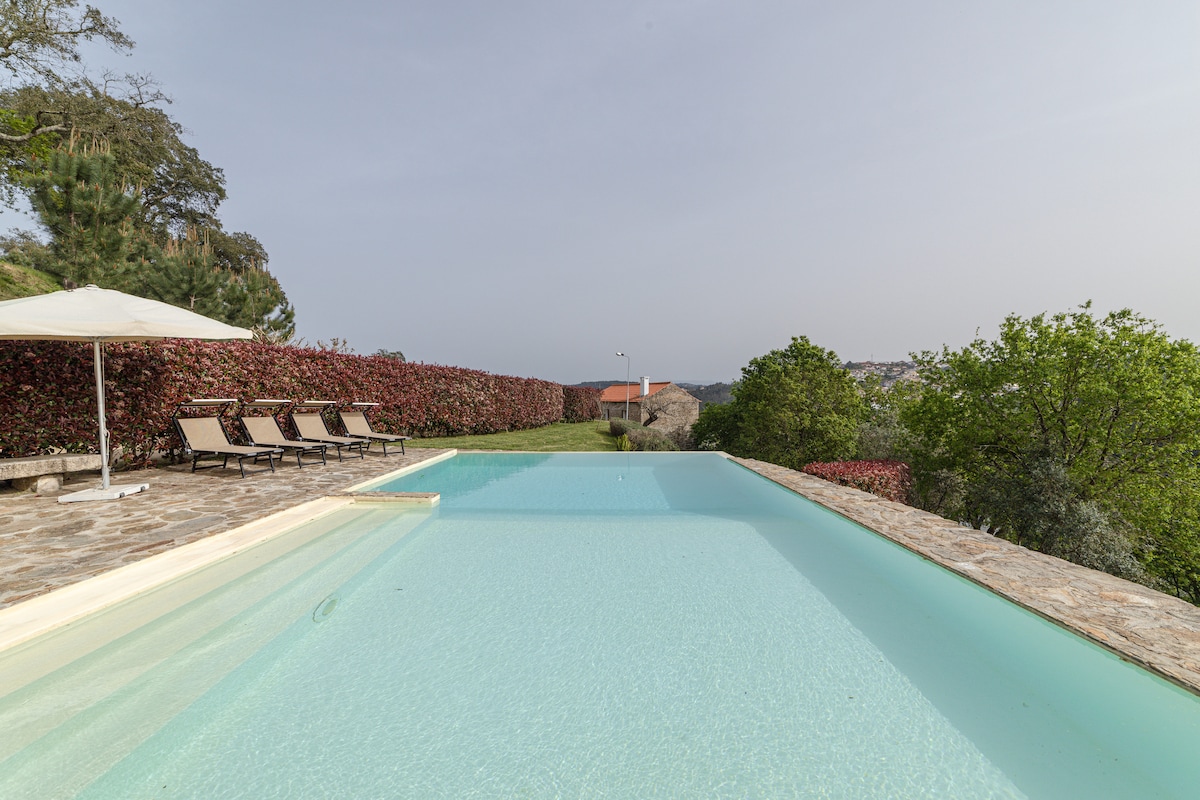 An infinity pool extends into the horizon, surrounded by a neatly landscaped lawn edged with vibrant hedges. Several sun loungers are arranged on the stone patio, offering a relaxing spot to enjoy the tranquil views of the surroundings.