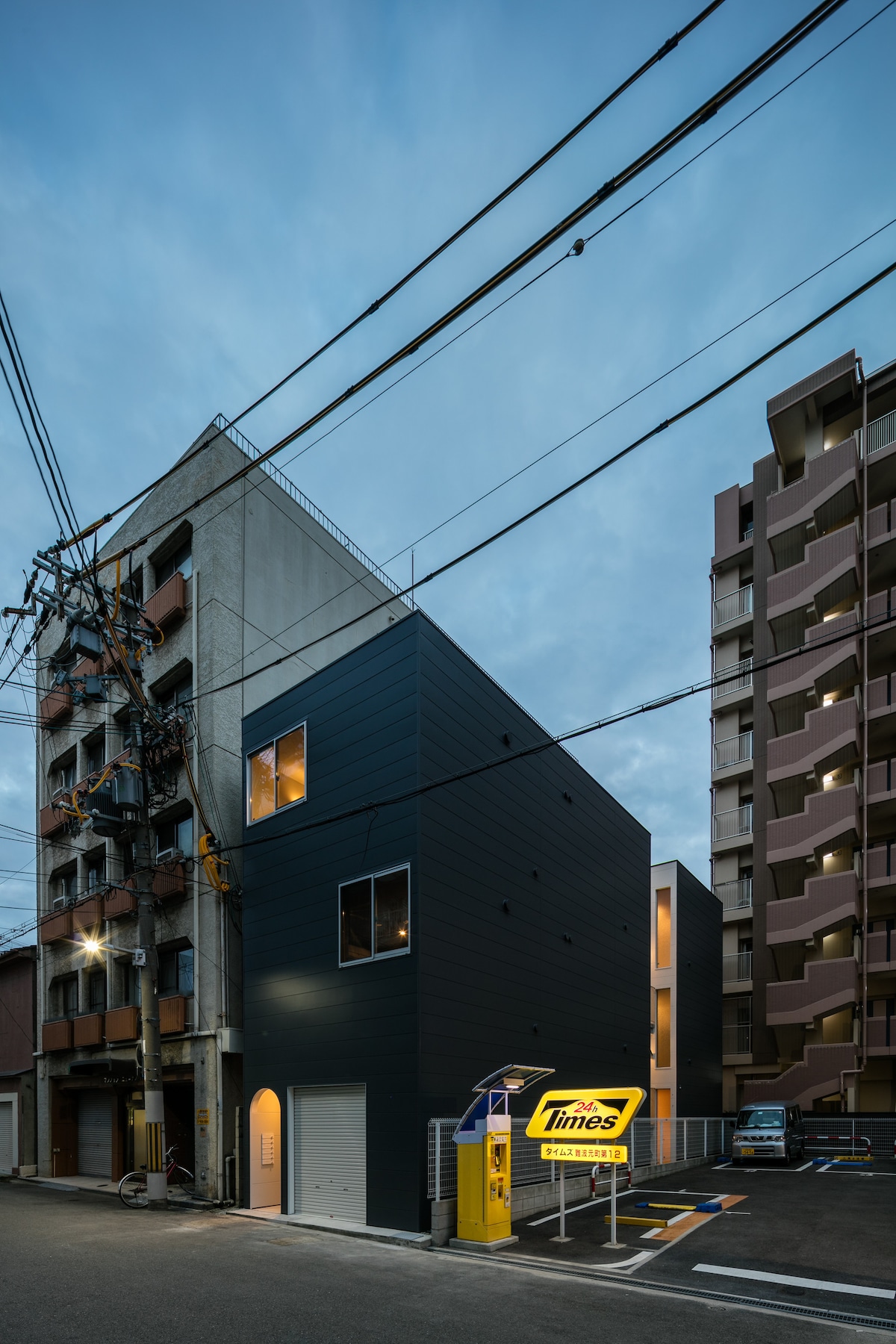 A modern building with a contrasting dark exterior faces the street, complemented by large windows that reflect the evening sky. Nearby structures feature varied architectural styles, and a brightly lit sign indicates a parking facility. Power lines run overhead, adding to the urban landscape.