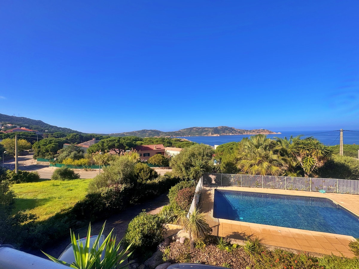 A view showcasing a serene coastal landscape with a clear blue sky and lush greenery. In the foreground, a secure swimming pool is framed by a spacious patio. The horizon reveals tranquil water and distant hills, enhancing the outdoor setting.