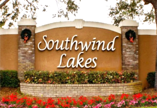 The Southwind Lakes entrance sign is prominently displayed against a backdrop of greenery. Seasonal wreaths adorn each side, adding a festive touch. Vibrant flowers border the base, enhancing the welcoming atmosphere of this lakefront community.