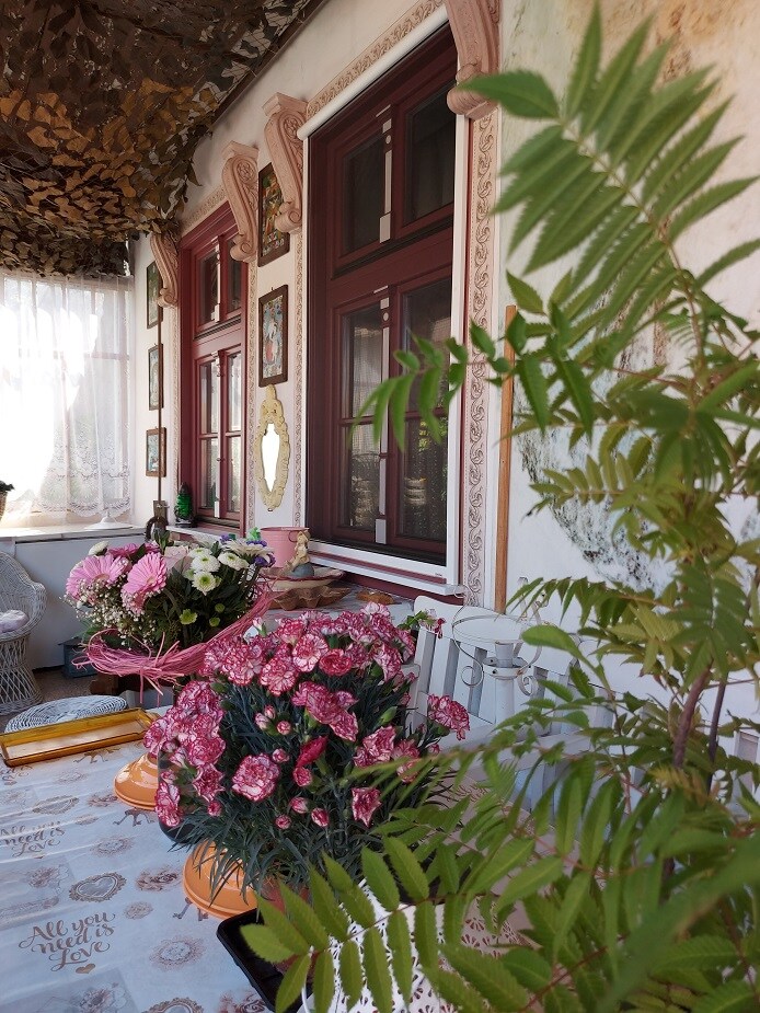 A table adorned with potted flowers and decorative items is positioned near large windows with red frames. Natural light filters through sheer curtains, illuminating the space. The surrounding walls are decorated with various framed art pieces, enhancing the inviting interior.