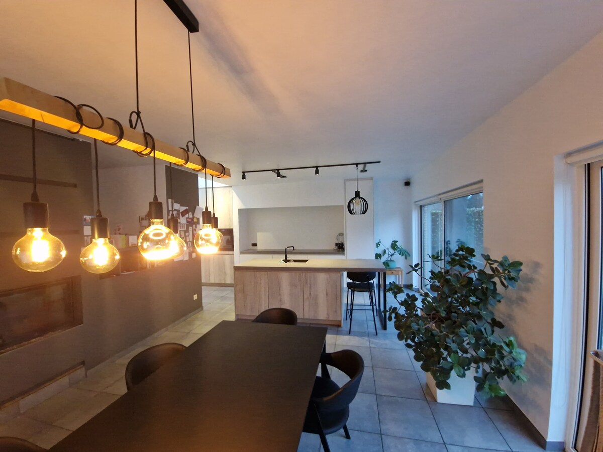 The dining area is highlighted by a long dark table surrounded by black chairs. Overhead, warm pendant lights hang, illuminating the space. A modern kitchen with a light wooden finish and a sink is visible in the background, along with a sizeable window that lets in natural light.