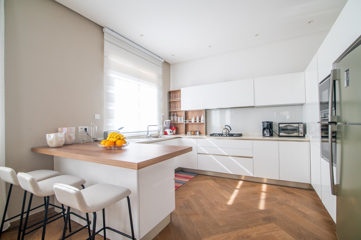 A modern kitchen is displayed with sleek white cabinetry and a spacious countertop. Three bar stools are positioned at the counter, while natural light fills the space through a window. Appliances including a microwave and refrigerator are visible, along with a bowl of fresh fruit.