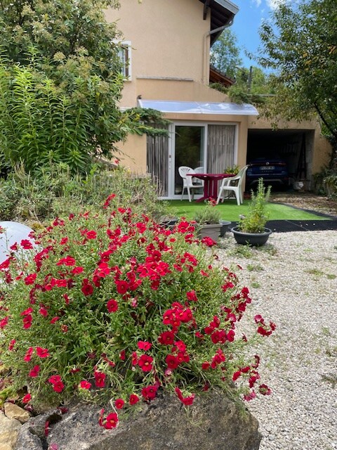 A charming exterior view of the villa is showcased, featuring vibrant red flowers in bloom. A shaded patio area with simple white chairs and a table offers a welcoming outdoor space adjacent to the property. Lush greenery surrounds the area, enhancing the natural ambiance.