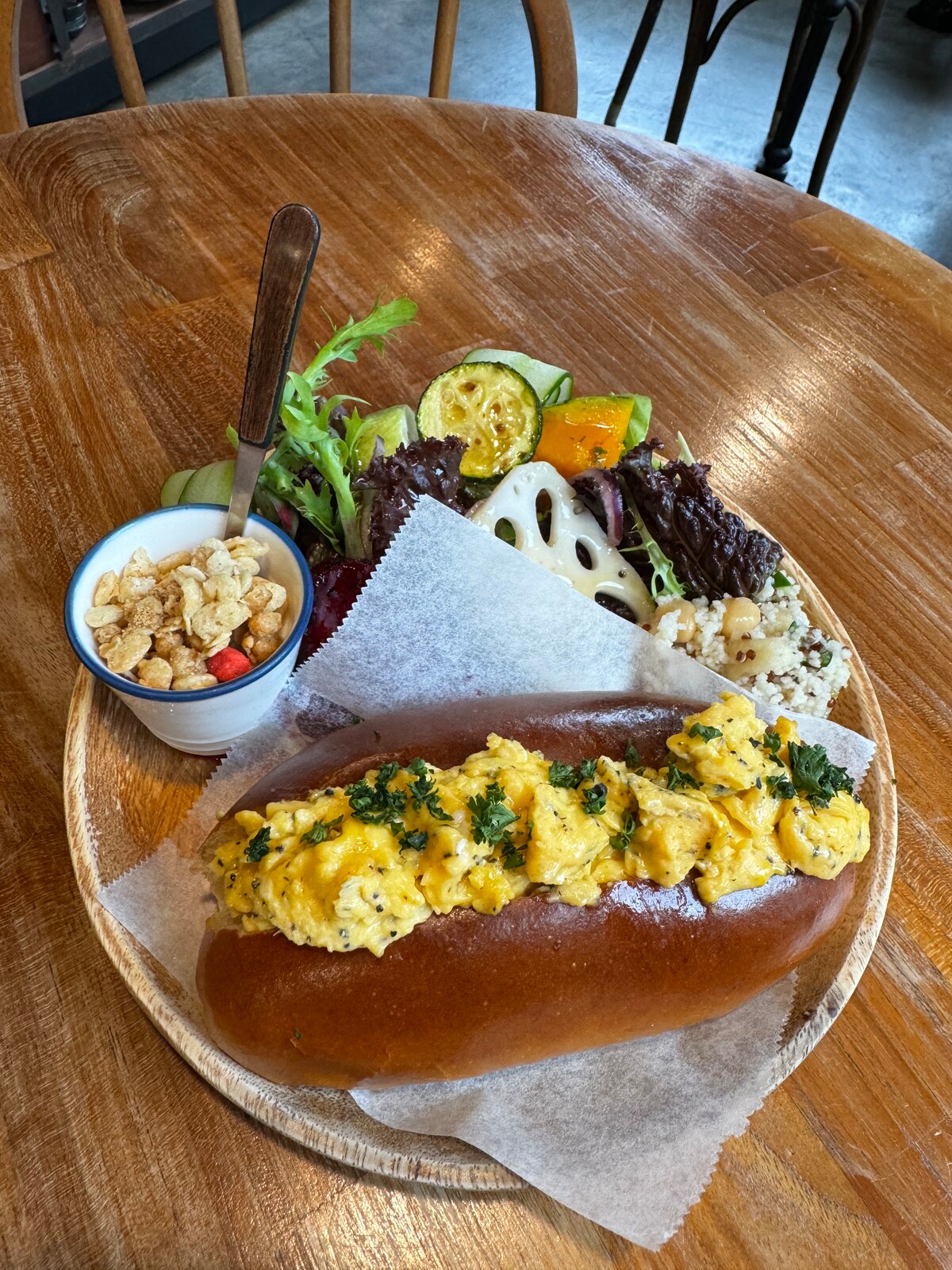 A wooden plate displays a golden-brown bun filled with fluffy scrambled eggs, garnished with fresh herbs. Accompanying the bun are a variety of colorful vegetables, including slices of zucchini and lettuce. A small bowl of crunchy snacks adds an enticing touch.