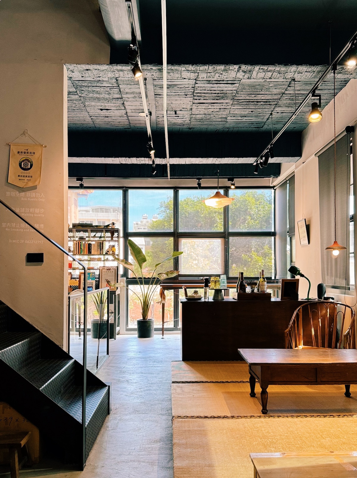 The entrance area features large windows allowing natural light to flood in, illuminating the spacious interior. A wooden table sits at the center, with surrounding seating. Plants are positioned nearby, enhancing the inviting atmosphere of the communal space.