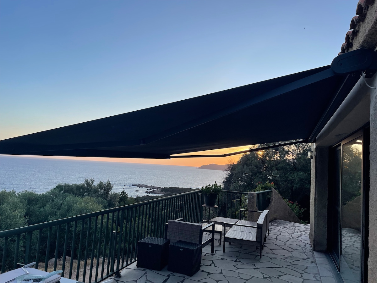A covered terrace is visible, featuring outdoor lounges and a dining area. The dark awning provides shade, while the view of the expansive coastline and sunset can be seen in the distance. The area is surrounded by greenery, contributing to a sense of tranquility.