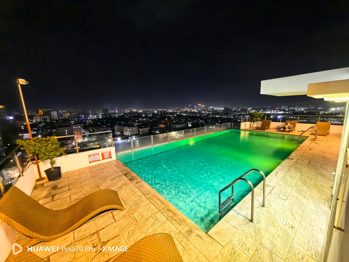 An infinity pool is viewed from a high vantage point, with water illuminated in green hues reflecting city lights at night. Lounge chairs are positioned nearby, offering a serene space for relaxation against a backdrop of the urban skyline.