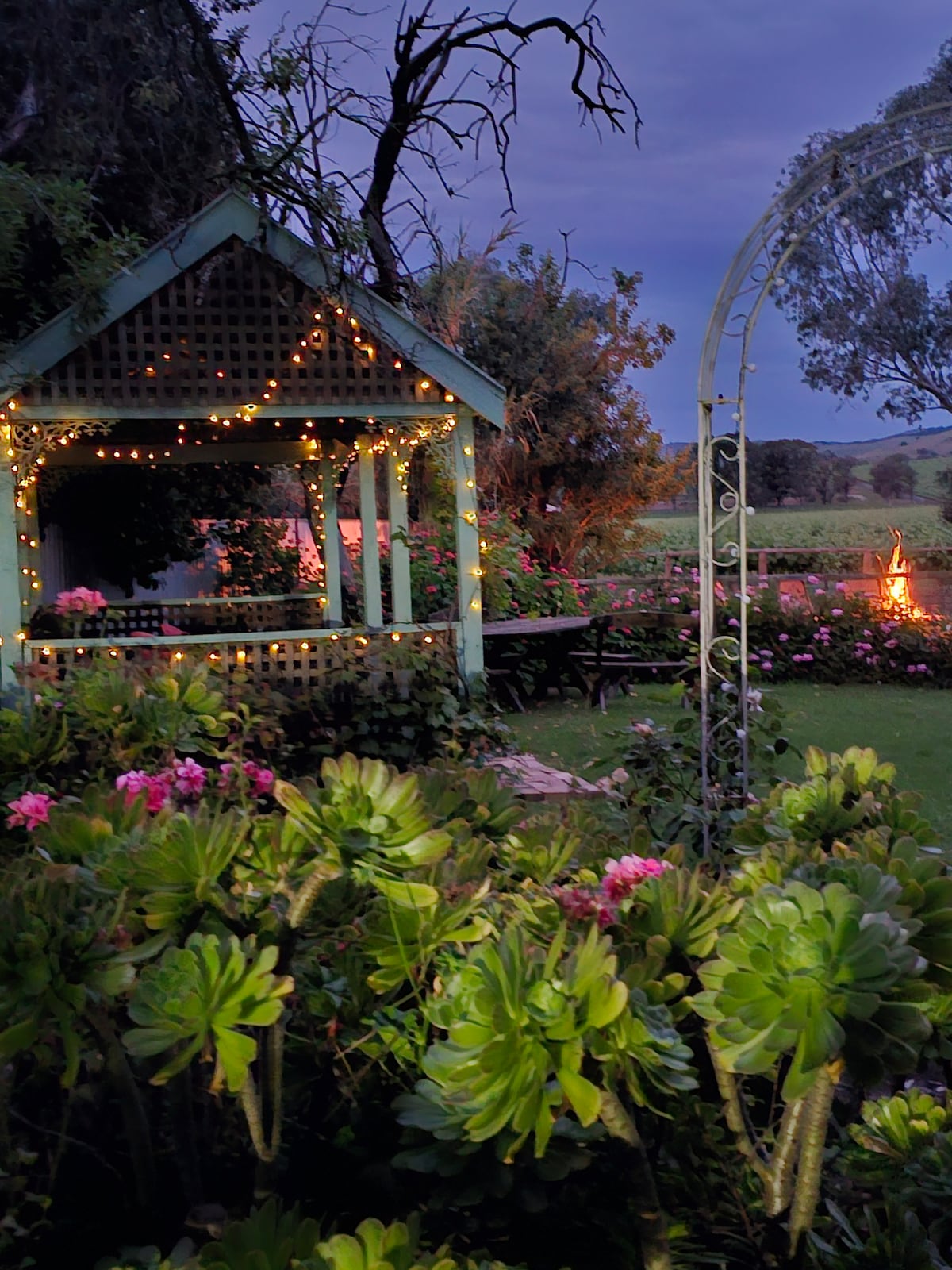 A charming gazebo adorned with string lights is framed by vibrant flower beds, creating a serene outdoor experience. A cozy fire pit is visible in the background, surrounded by lush greenery, contributing to a peaceful ambiance in the garden.