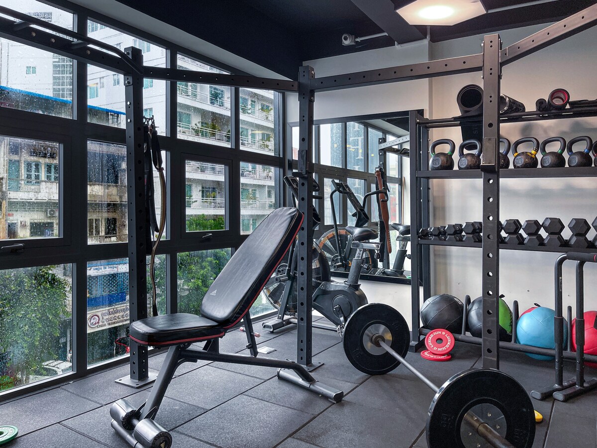 A well-equipped gym is highlighted by large windows that let in natural light. Various weights and kettlebells are neatly arranged alongside a bench press and exercise equipment, providing a functional space for workouts.