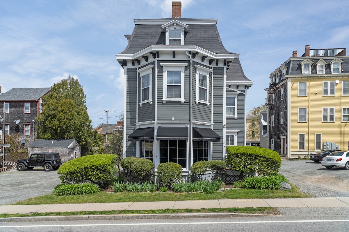 The exterior of a Victorian-style building is visible, characterized by its gray facade and distinctive architectural details. Lush green bushes frame the entrance, and multiple large windows allow natural light to enter. A shared parking area is situated in front, accommodating resident vehicles.