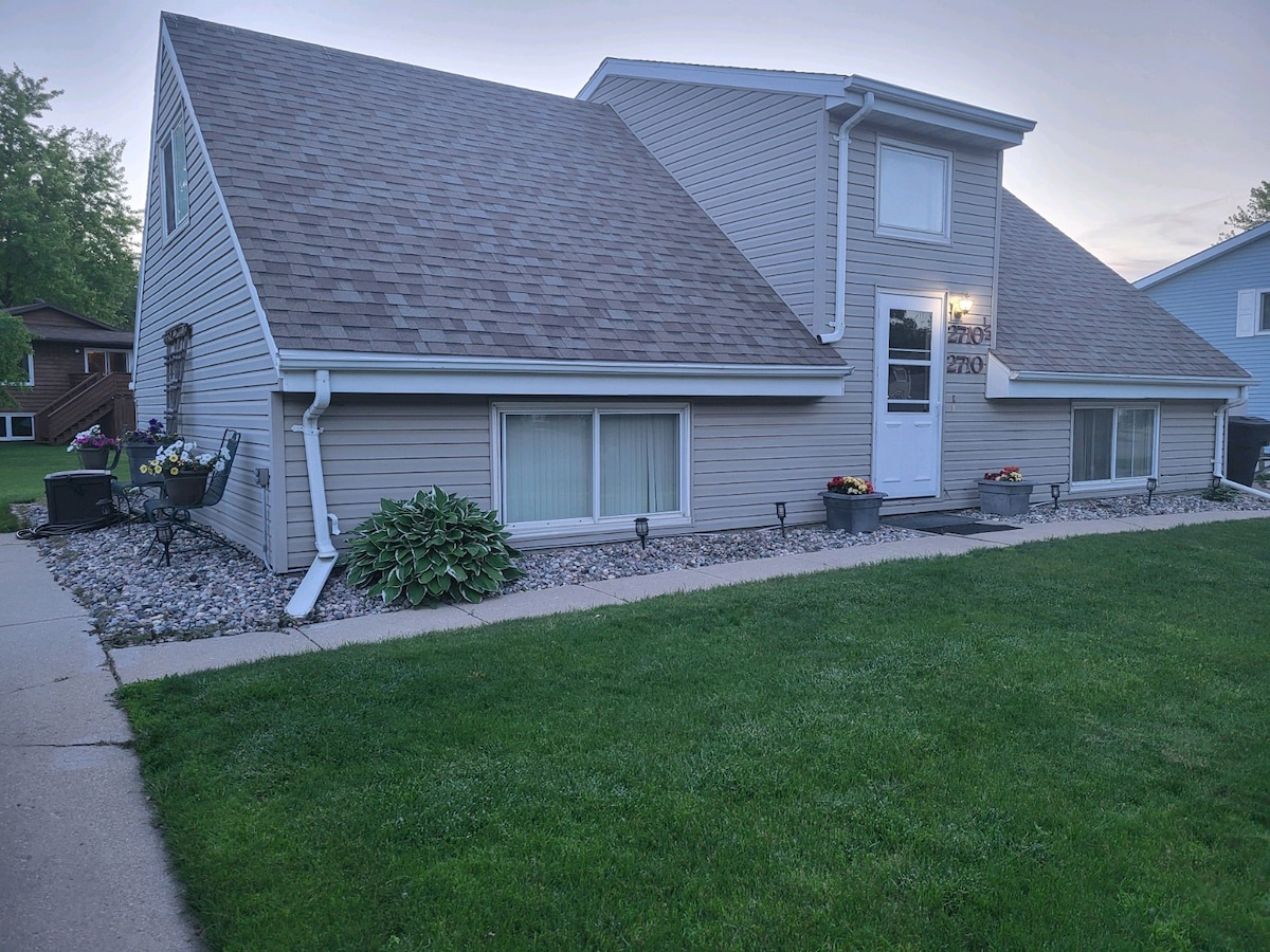A two-story duplex features a sloped roof and light grey siding. The front yard is landscaped with grass and various planters filled with blooms. Windows are positioned at the front, allowing natural light to fill the space. A concrete path leads to the entrance door.