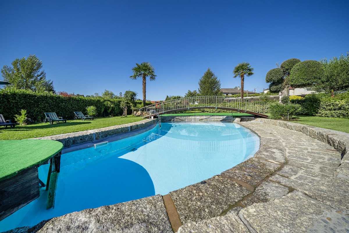 A clear, inviting pool is surrounded by lush greenery and palm trees. A stone bridge arches over the pool, connecting the landscaped garden areas. Comfortable lounge chairs are placed along the poolside, offering relaxation in a peaceful outdoor setting.