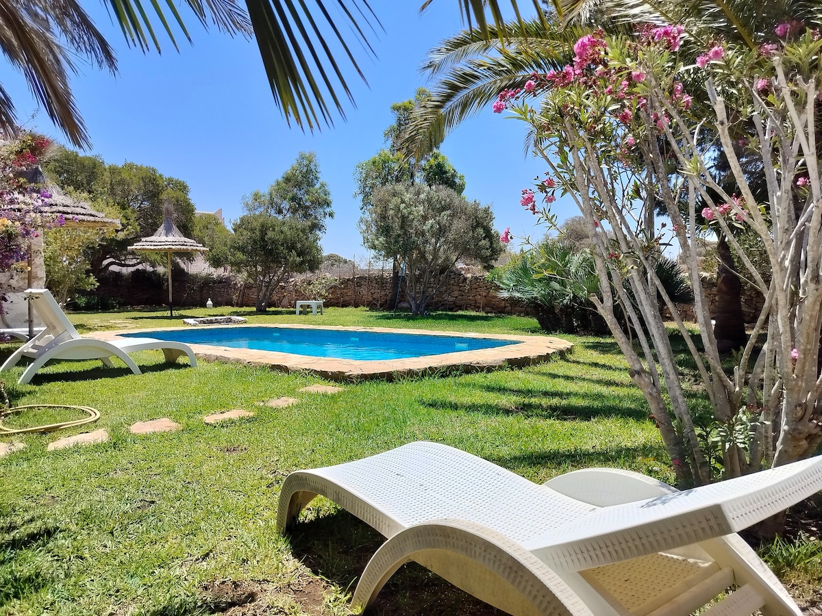 A tranquil garden area is depicted, featuring a private swimming pool surrounded by lush green grass. Sun loungers are positioned beside the pool, and palm trees along with flowering shrubs add a natural touch to the serene landscape.