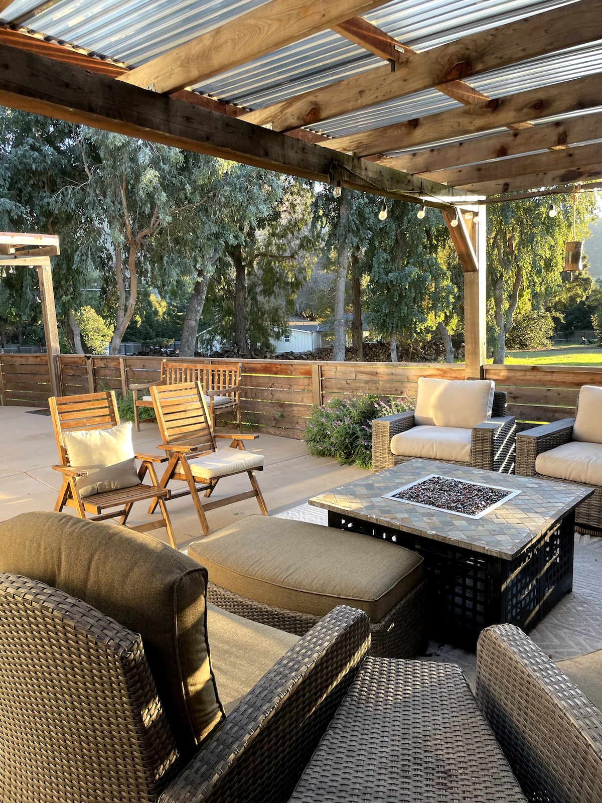 An outdoor patio area features comfortable seating with soft cushions arranged around a stone-topped coffee table. A fire pit is centered on the table, and a pergola adorned with lights casts a warm glow. Surrounding trees provide shade and privacy.
