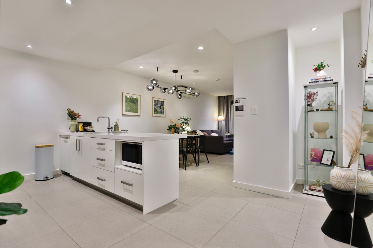 A sleek kitchen area is highlighted with a modern island counter featuring a microwave and storage drawers. Soft lighting illuminates the space, while vibrant plants and artwork add a touch of color. A dining table for four is visible in the adjoining living area.