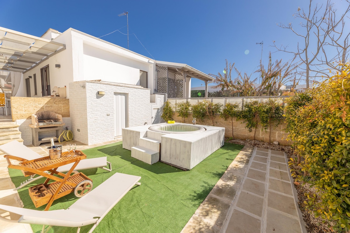 A private outdoor space features a hot tub surrounded by neatly arranged greenery and a well-maintained lawn. Sun loungers offer a place to relax, while a barbecue grill is positioned for outdoor cooking. Clear blue skies are visible overhead, contributing to the serene ambiance.