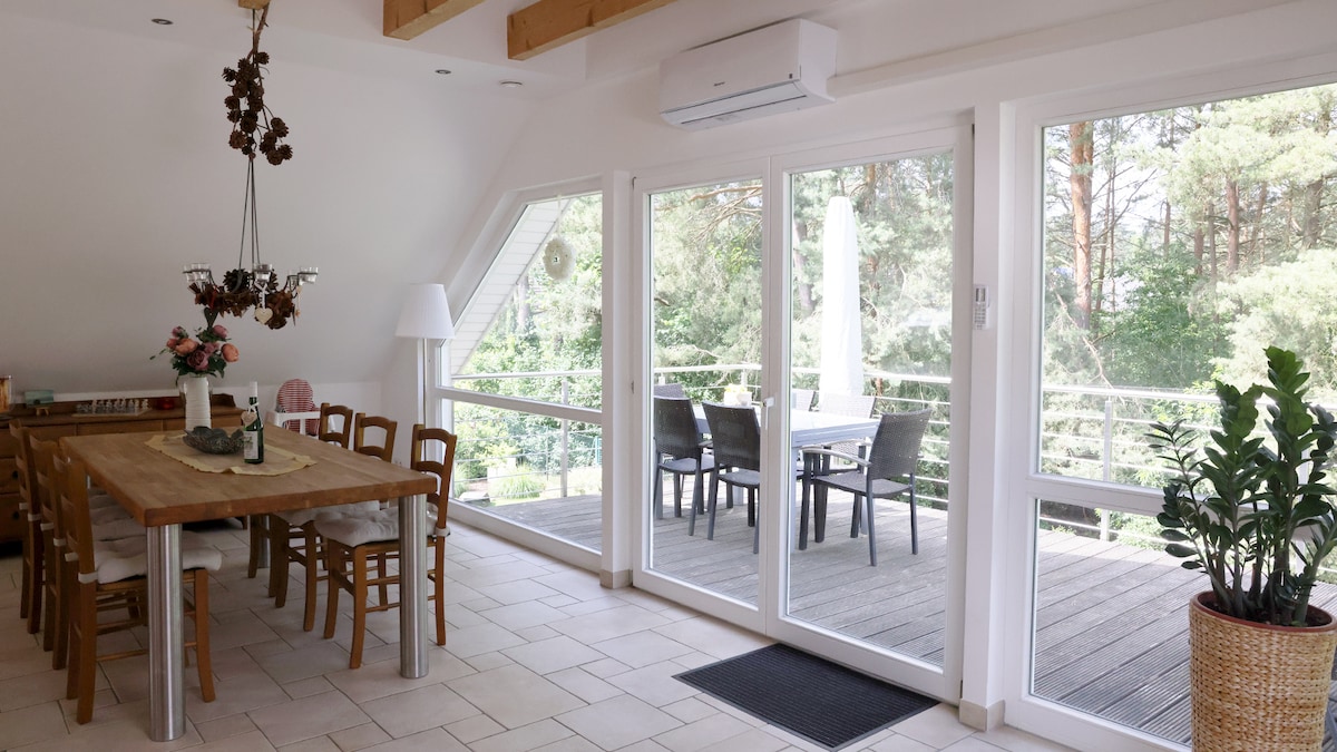 A bright dining area is featured, with a large wooden table surrounded by several chairs. Floor-to-ceiling windows provide views of a serene wooded landscape. A balcony with outdoor seating is visible, complemented by greenery and natural light flowing into the space.