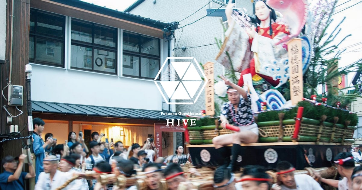 A lively street scene captures a traditional festival procession. A decorated float is prominently displayed, surrounded by a crowd of participants. Nearby buildings, including Fukuoka Guesthouse HIVE, provide a backdrop to the festive atmosphere.
