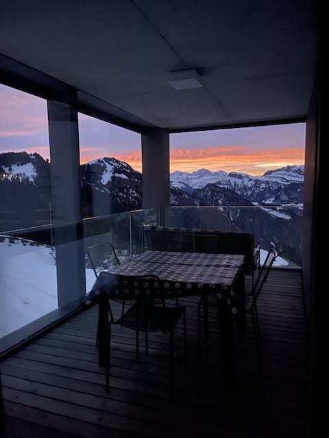 A cozy balcony with a wooden floor features a dining table surrounded by several chairs. Large glass panels frame a stunning view of snow-capped mountains, while a vibrant sunset casts warm colors across the sky. The serene atmosphere is enhanced by the surrounding nature.