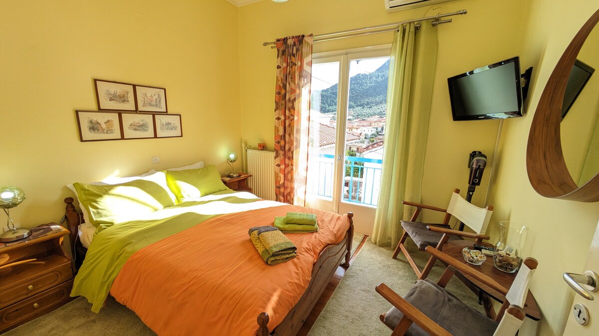 The bright bedroom features a spacious bed adorned with vibrant orange and green bedding. A balcony door invites natural light, revealing picturesque views. Two bedside lamps provide soft illumination, and a television is mounted on the wall. A seating area includes a wooden chair and a small table.
