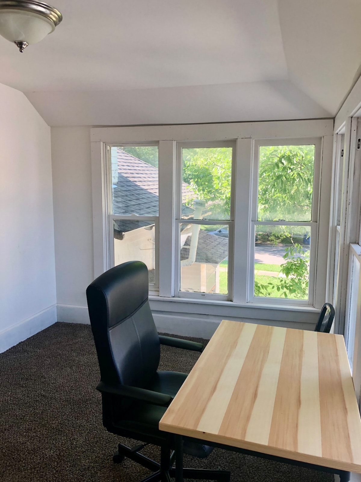 A corner workspace is depicted, featuring a wooden desk paired with a black ergonomic chair. Sunlight enters through a set of four large windows, illuminating the room and providing a view of the outside greenery.