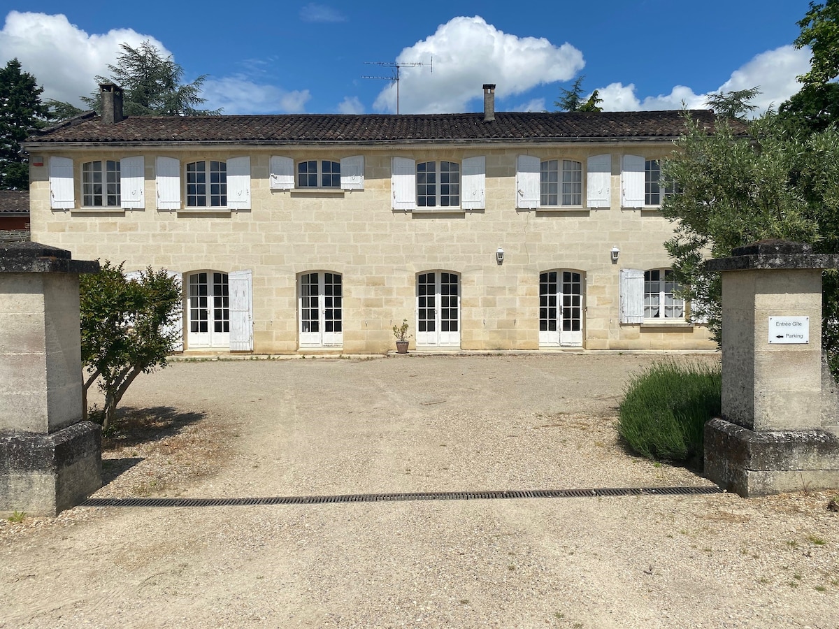 The exterior of the 19th-century residence is visible, featuring a stone facade with white shutters on multiple windows. A gravel driveway leads to the entrance, flanked by simple stone pillars. Greenery surrounds the property, and blue skies with scattered clouds are present above.