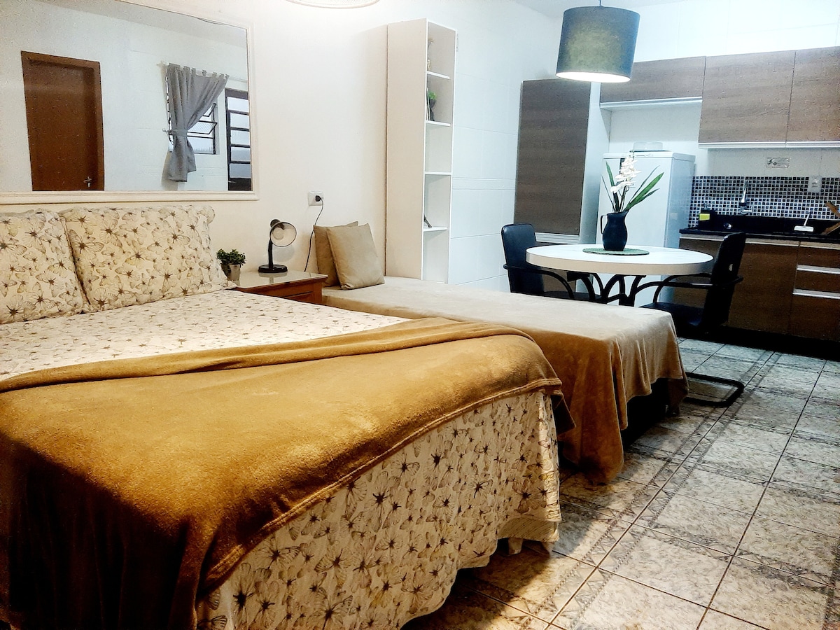 A compact suite features a spacious bed with a soft throw and a cozy second bed. A small dining area is provided with a round table and chairs, while the kitchenette is equipped with modern appliances, all set within an open layout.