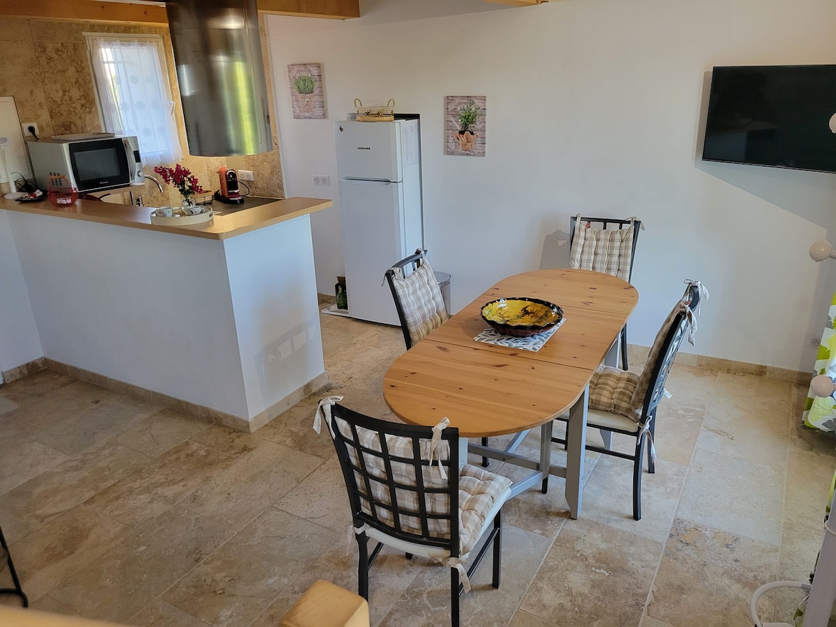 A dining area is presented with a wooden oval table surrounded by six metal chairs with cushions. A kitchenette is visible in the background, featuring a refrigerator and a coffee maker. Soft lighting illuminates the space, enhancing the neutral tones of the stone floor.