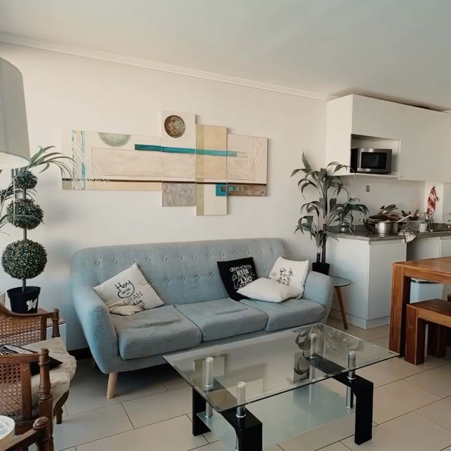 A bright living area features a light grey sofa against a beige wall with abstract artwork. A glass coffee table sits atop tiled flooring, surrounded by a wooden dining table and chairs. Green plants provide a touch of nature, while a small side table adds functionality.