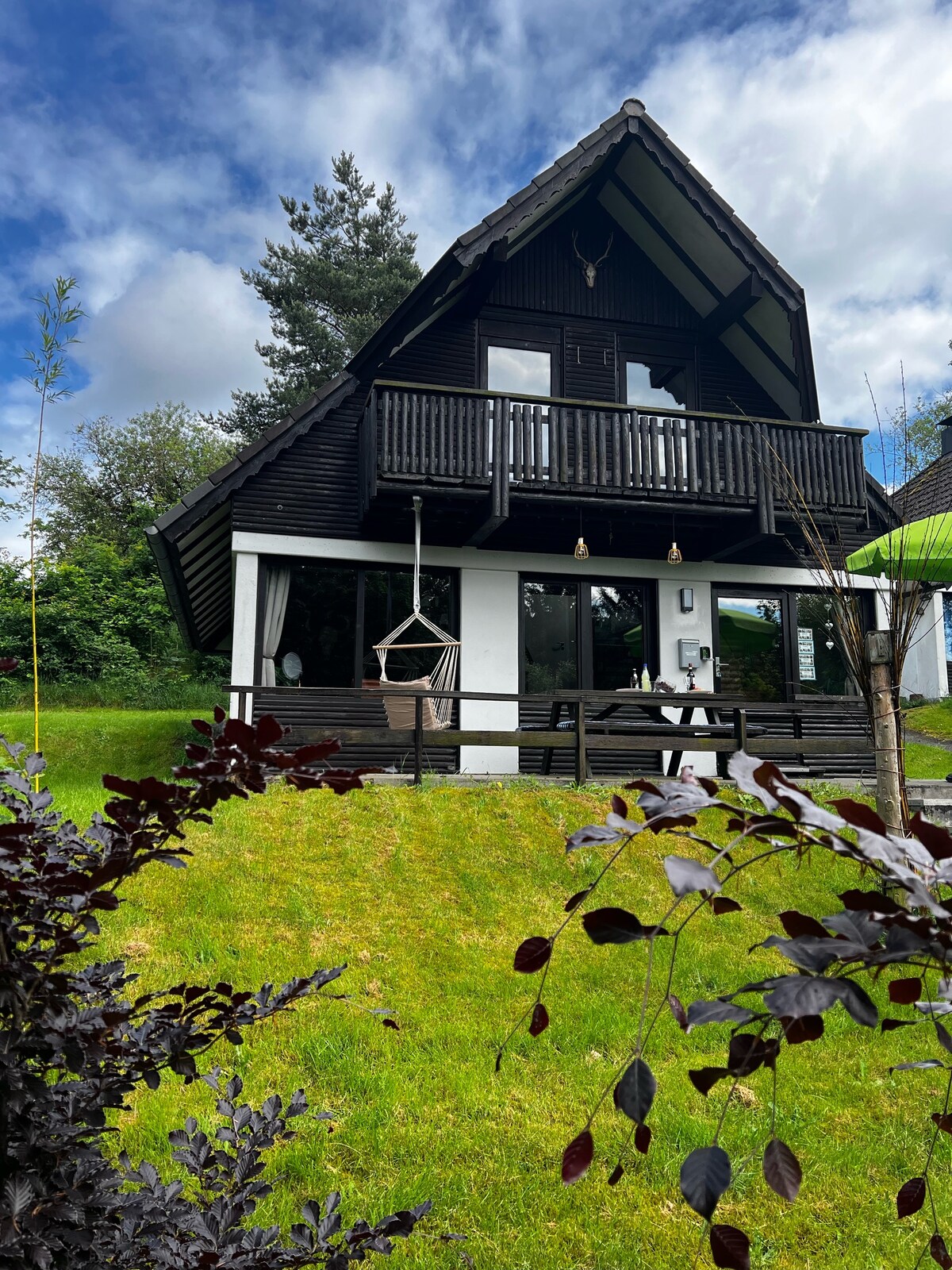 A three-story wooden house is set against a backdrop of greenery. The structure features a balcony with a hammock and large glass doors below, allowing for natural light. The surrounding landscape includes a well-maintained lawn with various shrubs and trees.