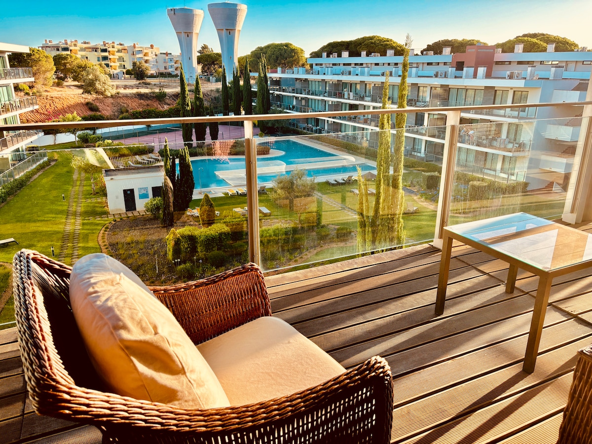 A private balcony features a comfortable wicker chair with a plush cushion. The view showcases a landscaped area with vibrant greenery and a large swimming pool. Modern buildings form a backdrop under soft evening light, creating a serene outdoor setting.