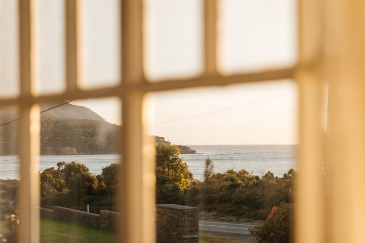 A view from an antique window frames a serene landscape, featuring rolling hills meeting the ocean. The scene captures the gentle waves and coastal vegetation in the warm light of the setting sun.