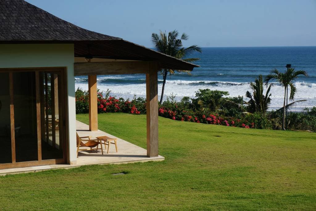 A spacious outdoor area features a manicured lawn overlooking the ocean. The seating area includes wooden chairs positioned on a covered patio, with vibrant flowering plants adding color to the landscape. The waves of the Indian Ocean create a scenic backdrop under a clear blue sky.
