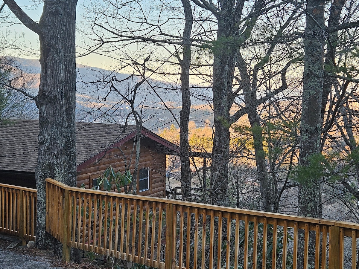 A view from the cabin shows a wooden fence and the surrounding trees, with glimpses of mountains in the distance. The setting sun casts a warm light, creating a serene atmosphere among the natural landscape.