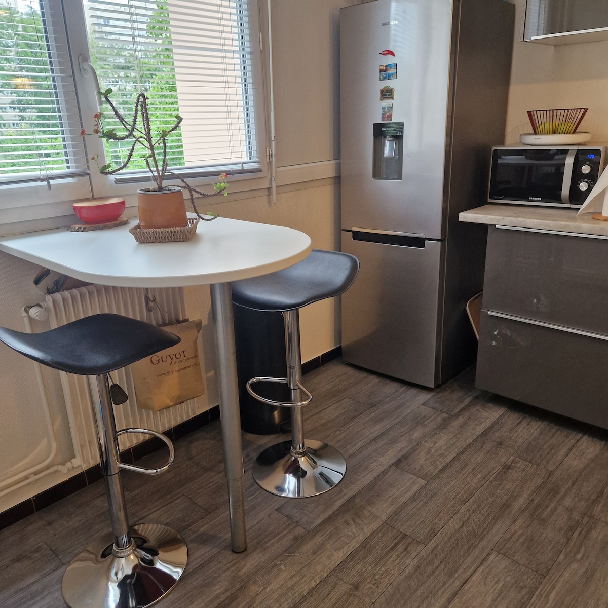 A compact kitchen area features a round white table with two sleek black stools. A silver refrigerator stands beside a microwave, complemented by a small decorative plant on the table. Natural light enters through the window, highlighting the modern cabinetry and neutral tones of the space.