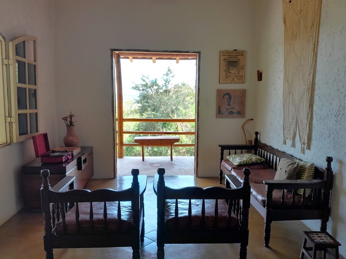 A sitting area is featured with two wooden benches and a small table, oriented towards a large opening leading to a balcony. Natural light filters in, highlighting the simplicity of the decor and the view of surrounding greenery.