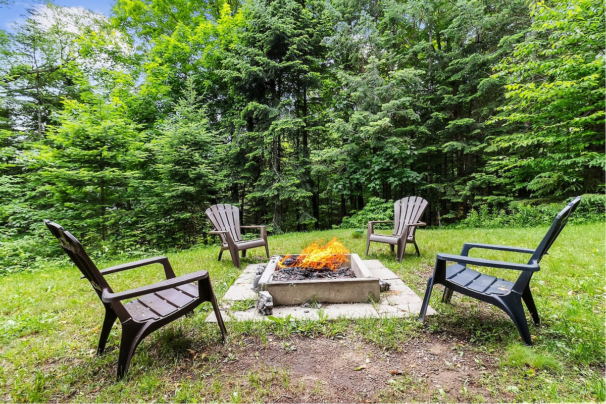A circular firepit area is set within a lush green landscape. Four wooden Adirondack chairs are positioned around the firepit, which features a flame flickering above a stone base. Tall trees in the background provide a natural, secluded atmosphere.