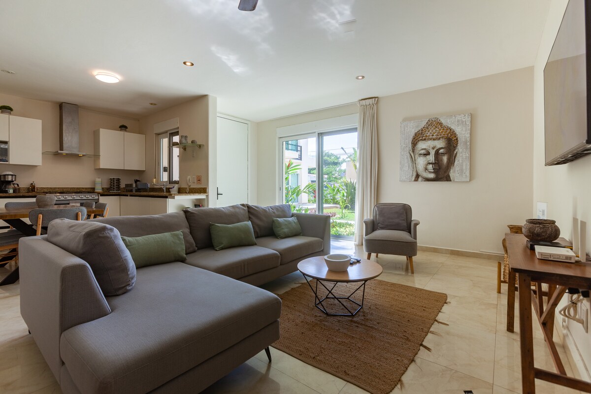 The living area features a spacious gray sofa, accented with green cushions, and is arranged around a wooden coffee table. Natural light floods in through large sliding glass doors that provide views of the garden. A modern wall art piece adds visual interest.